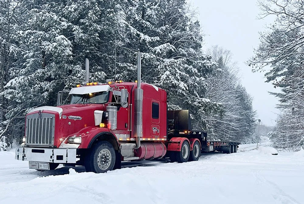 A red semi-truck with a flatbed trailer driving through a snow-covered road surrounded by snow-laden trees.