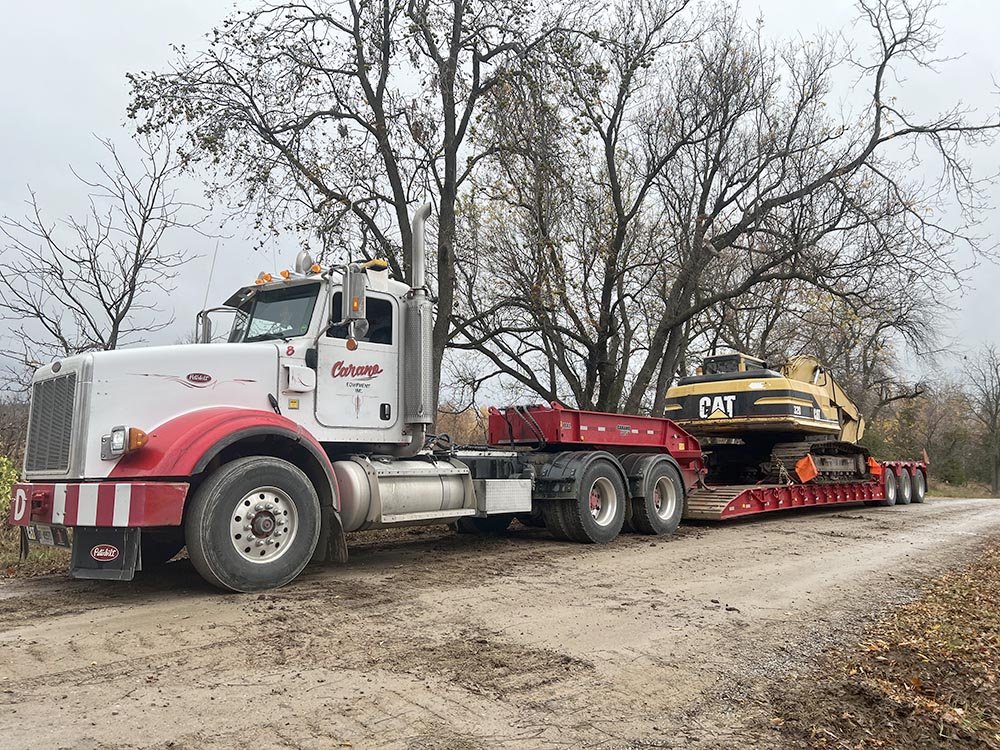 A semi-truck with a red and white cab parked on a dirt road, carrying a large yellow CAT excavator on a lowboy trailer, with leafless trees in the background.