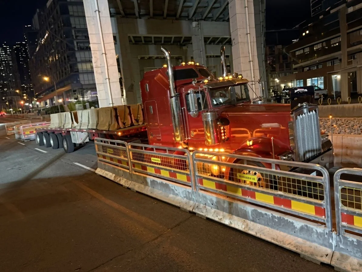 Red semi-truck parked on a city street at night, surrounded by construction barriers and fencing, with buildings and streetlights in the background.