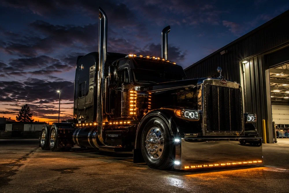 Black semi-truck with reflective chrome details parked outside a large warehouse at sunset.