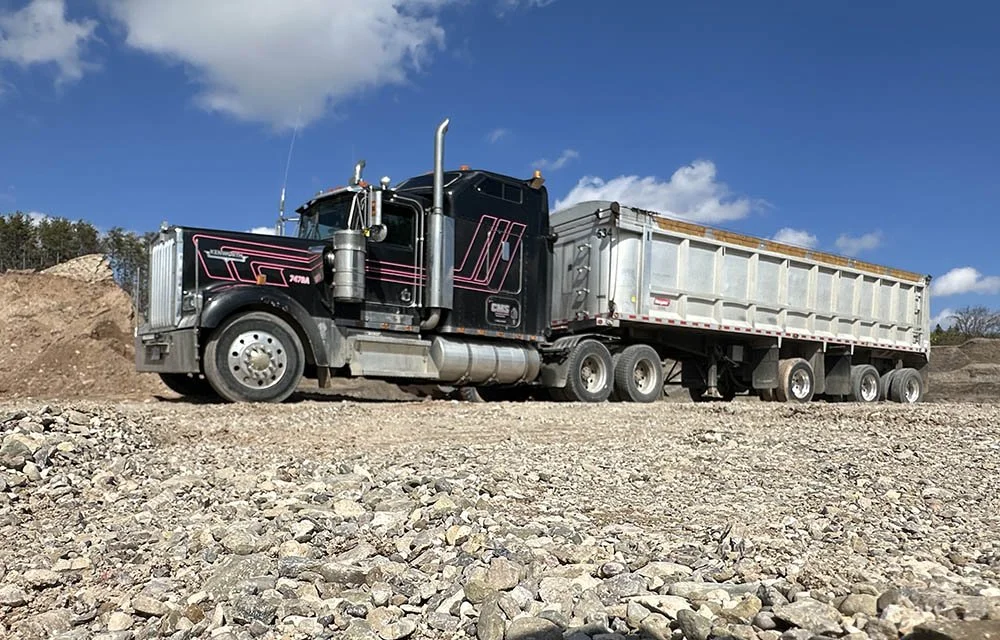 A large black semi-truck with a silver trailer loaded with dirt or gravel at a construction or mining site, under a partly cloudy blue sky.