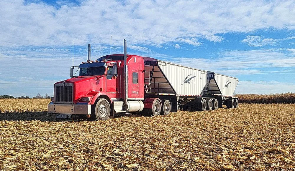 A red semi-truck with two large white cargo trailers parked in a harvested cornfield under a partly cloudy sky.