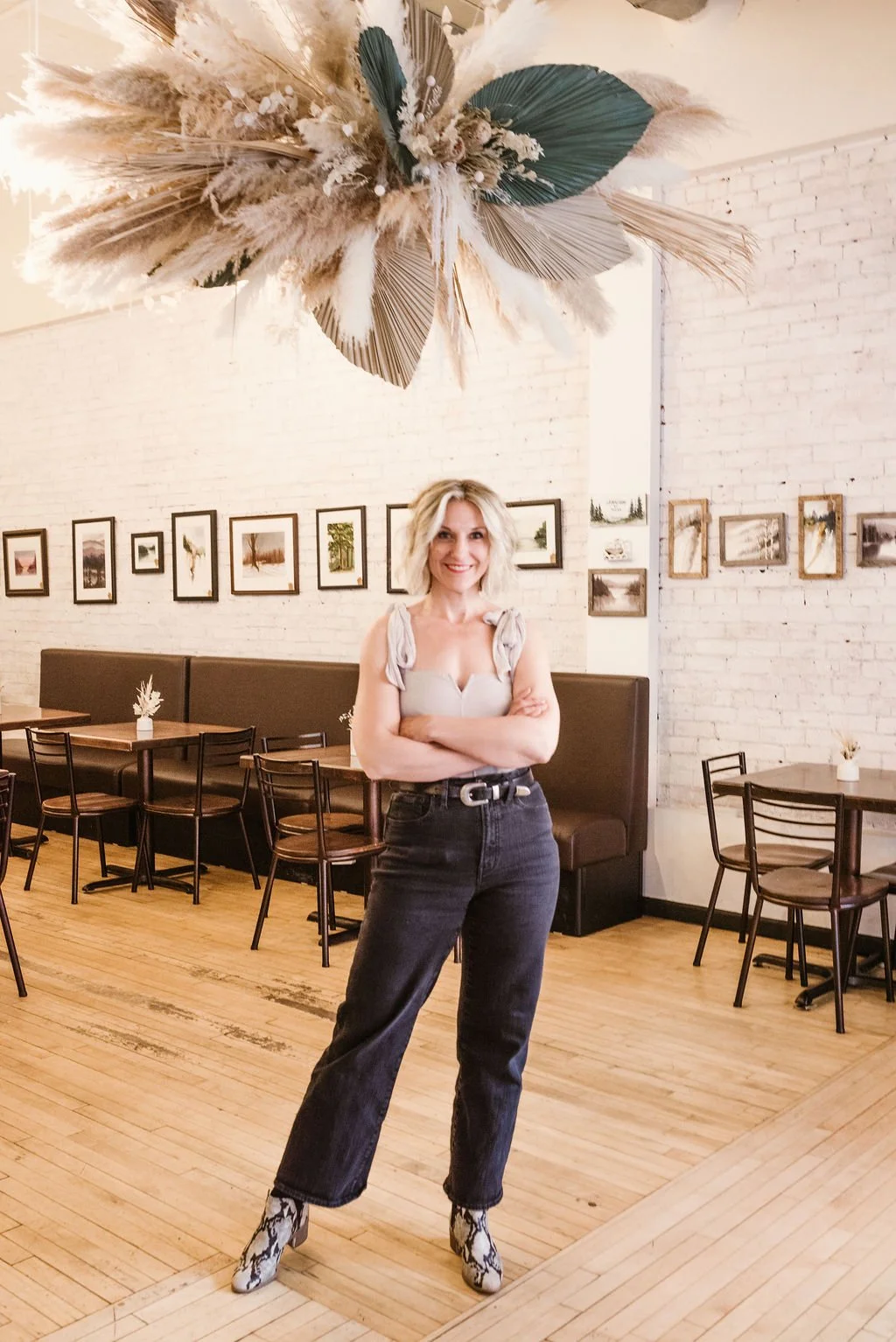 A woman with blonde hair, wearing a light-colored top, black jeans, and snake-patterned boots, stands with arms crossed in a modern cafe or restaurant with white brick walls, framed pictures, and a large hanging dried floral arrangement.
