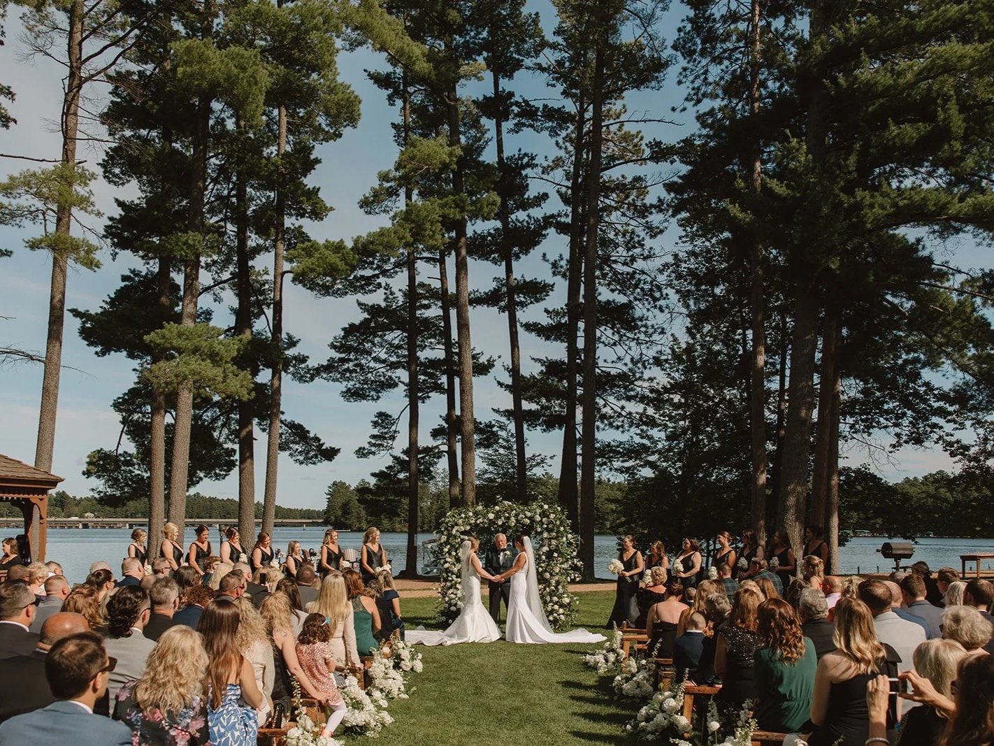 Outdoor wedding ceremony by a lake with tall trees. Bride and groom holding hands, officiant in the center. Bridesmaids and groomsmen standing on either side, guests seated facing the couple. Floral arrangements along the aisle.