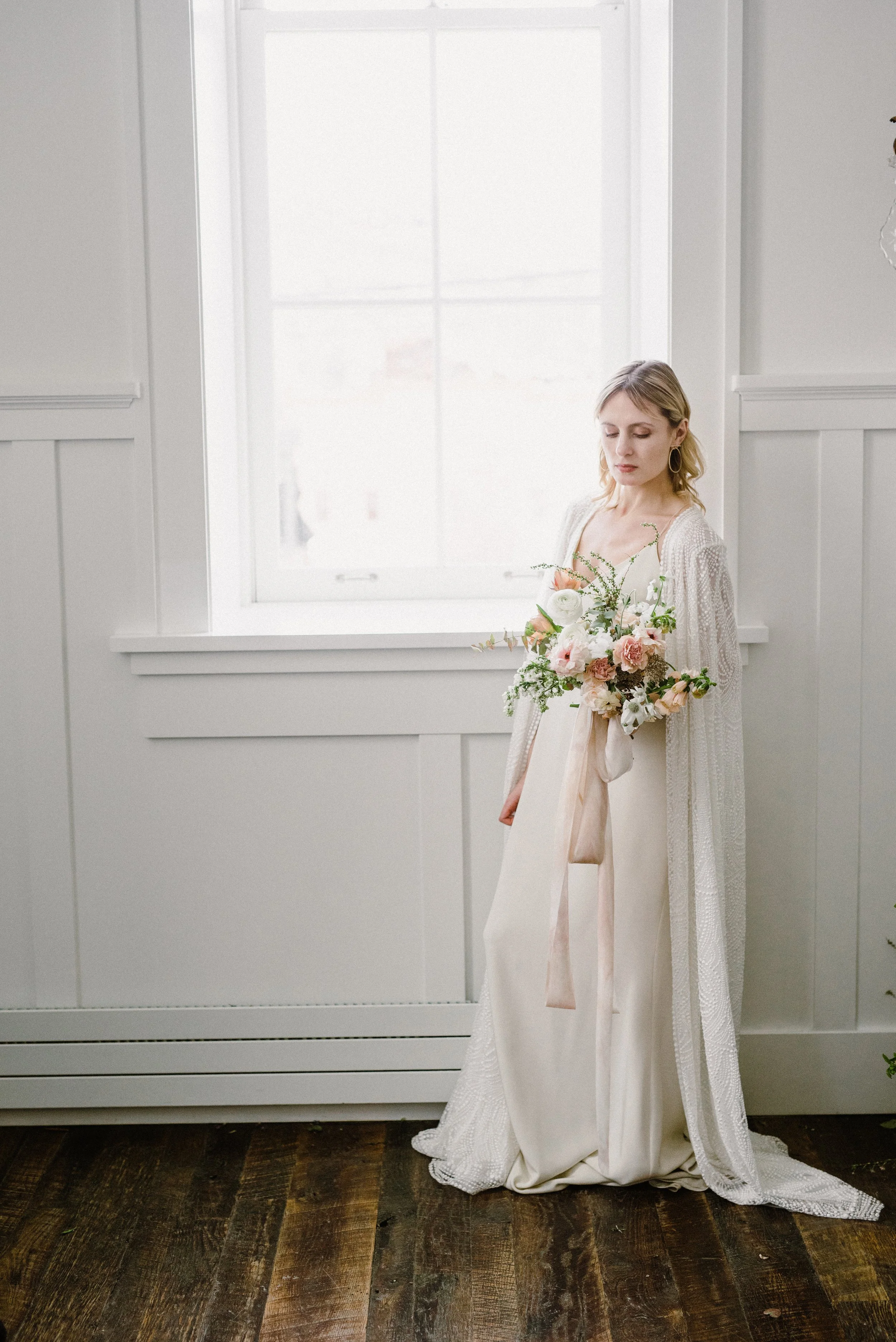 A woman in a white dress with a lace overlay standing by a white window holding a bouquet of light pink and white flowers. The room has white walls and dark wooden floor.