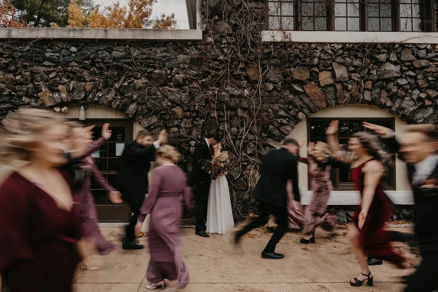A wedding celebration with a bride and groom standing against a stone wall while guests dance and celebrate in front of them.