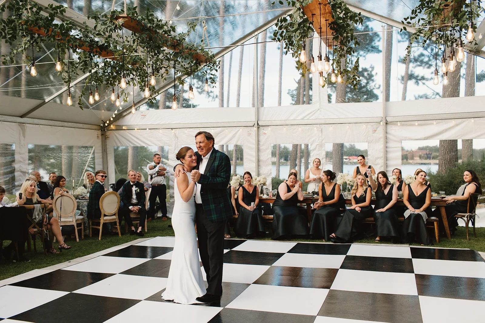 A bride and an older man, likely her father, dance together on a black-and-white checkered dance floor at a wedding reception inside a large white tent, with guests seated around watching.