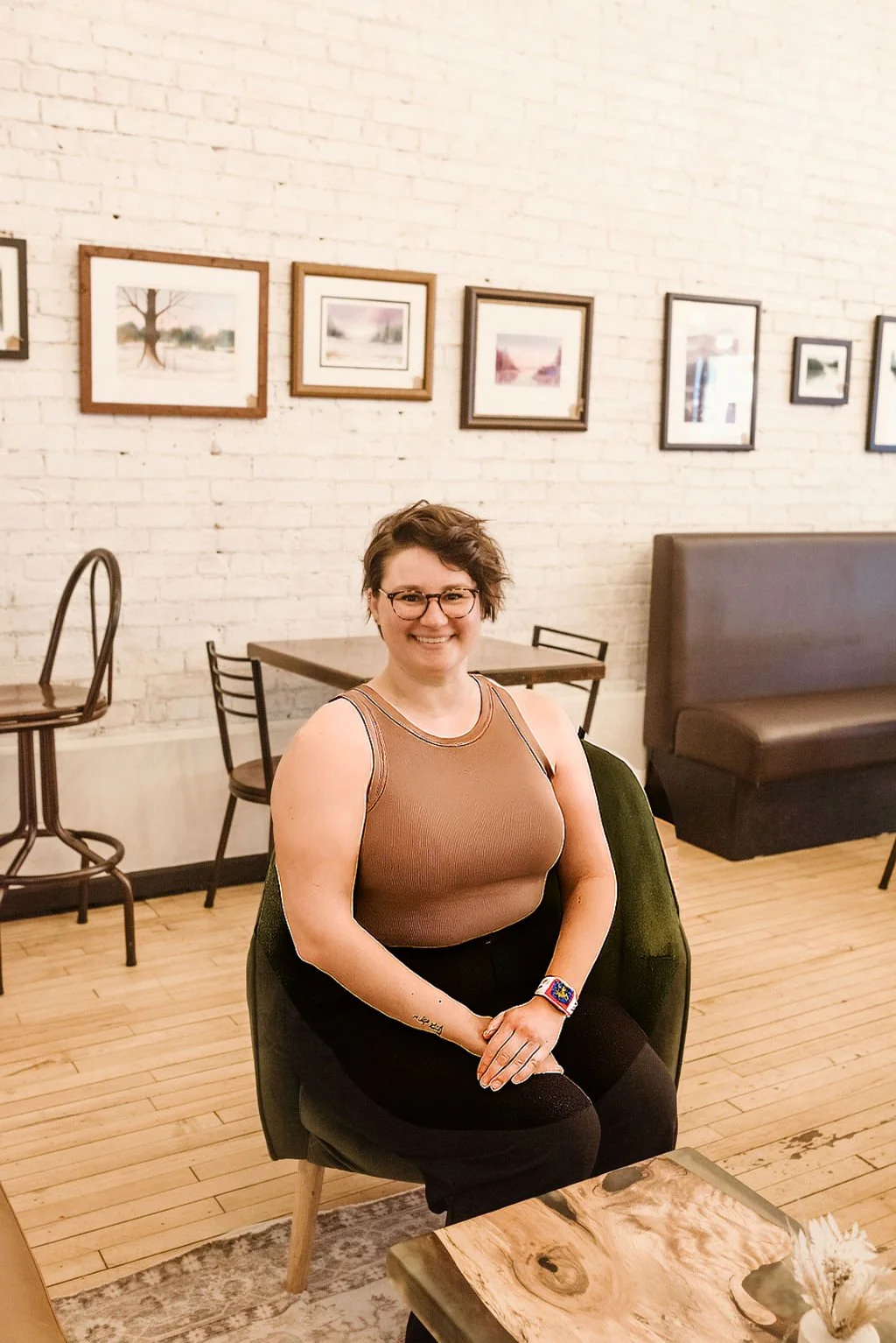A woman with short brown hair, glasses, and a tan sleeveless top, sitting on a green chair in a cafe with a white brick wall decorated with framed artwork behind her.