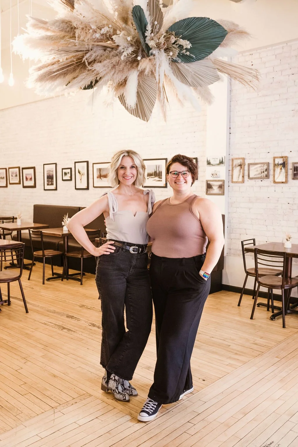 Two women standing inside a cafe, smiling at the camera. The woman on the left has blonde, wavy hair and is wearing a light-colored top with tie straps and black jeans. The woman on the right has short, dark hair, glasses, and is wearing a sleeveless brown top with black pants. The cafe has a white brick wall, framed pictures, wooden floors, and tables with chairs.
