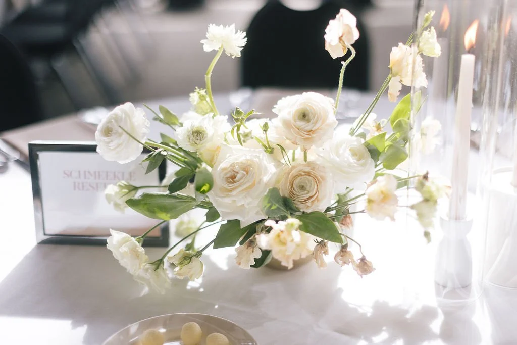 A white floral centerpiece with roses and greenery on a table at a formal event.