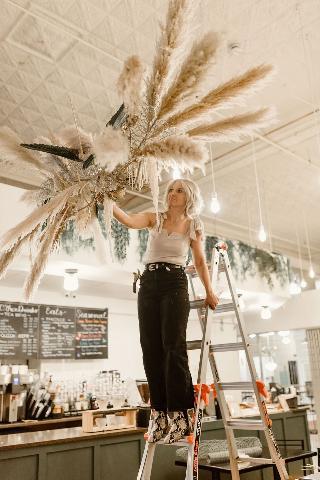 A woman standing on a ladder arranging a large dried flower arrangement in a cafe or restaurant setting.