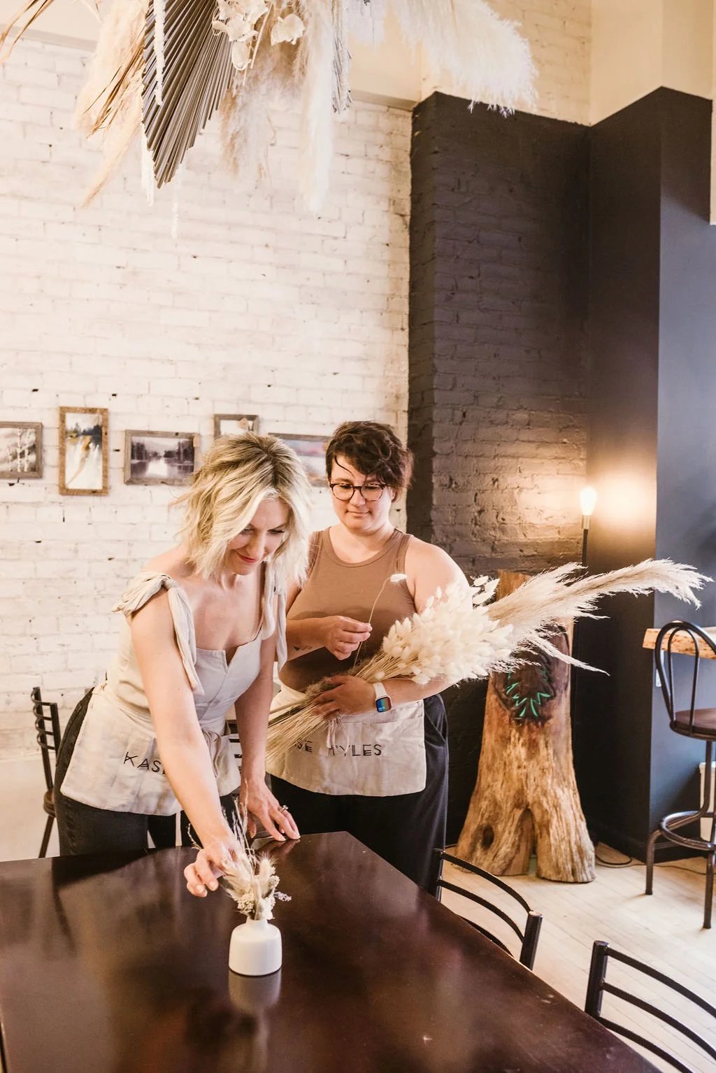 Two women preparing floral arrangements with dried pampas grass in a modern, cozy restaurant or cafe with white brick walls and minimal decor.