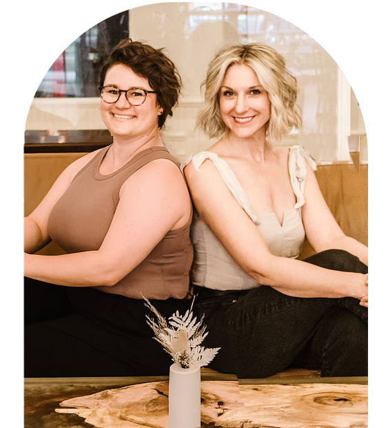 Two women sitting on a bench back-to-back in a restaurant, smiling at the camera, with a small bouquet of dried flowers on the table in front of them.