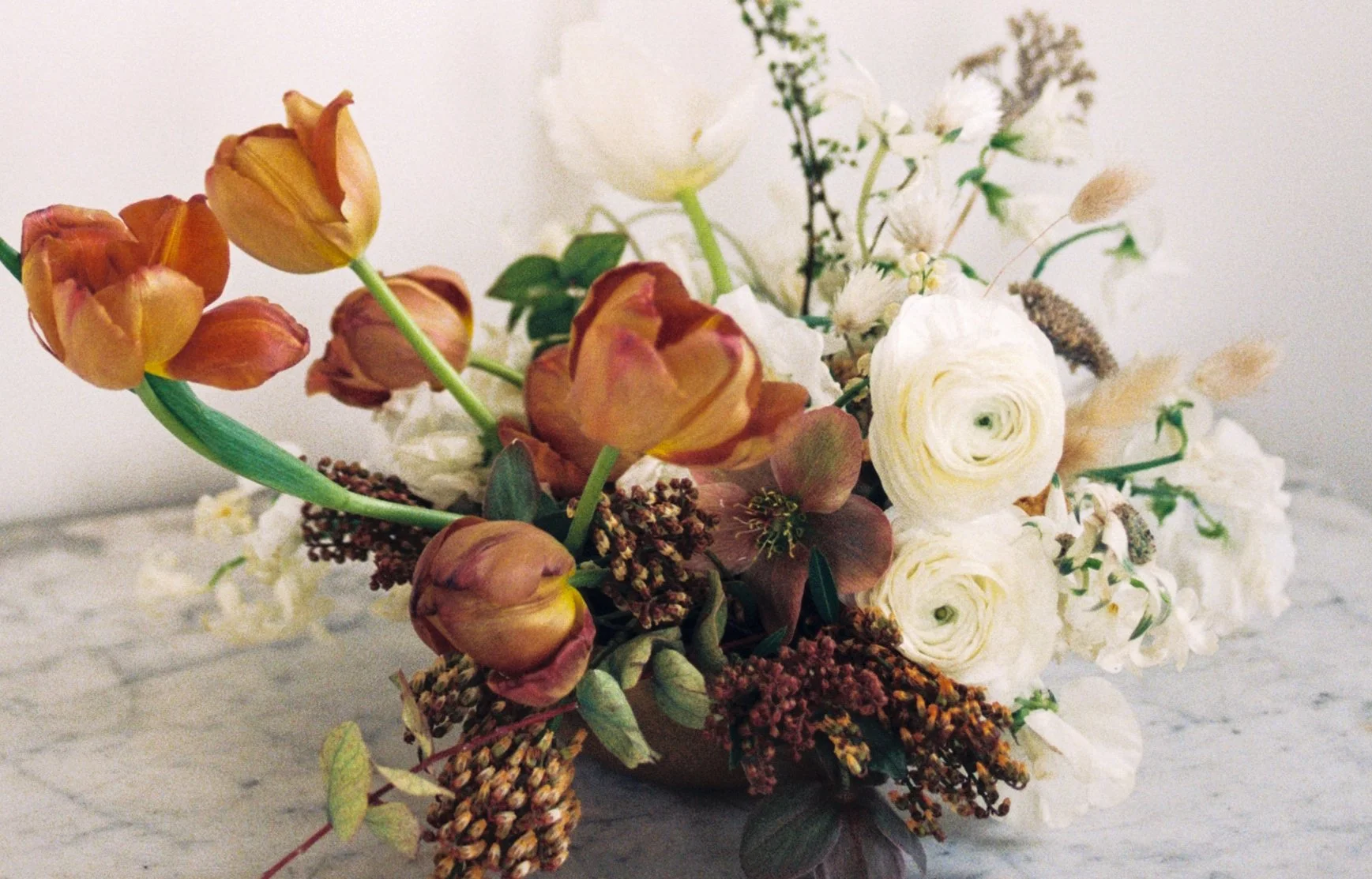 A bouquet of flowers featuring white, orange, and pink blossoms with green leaves in a brown vase.