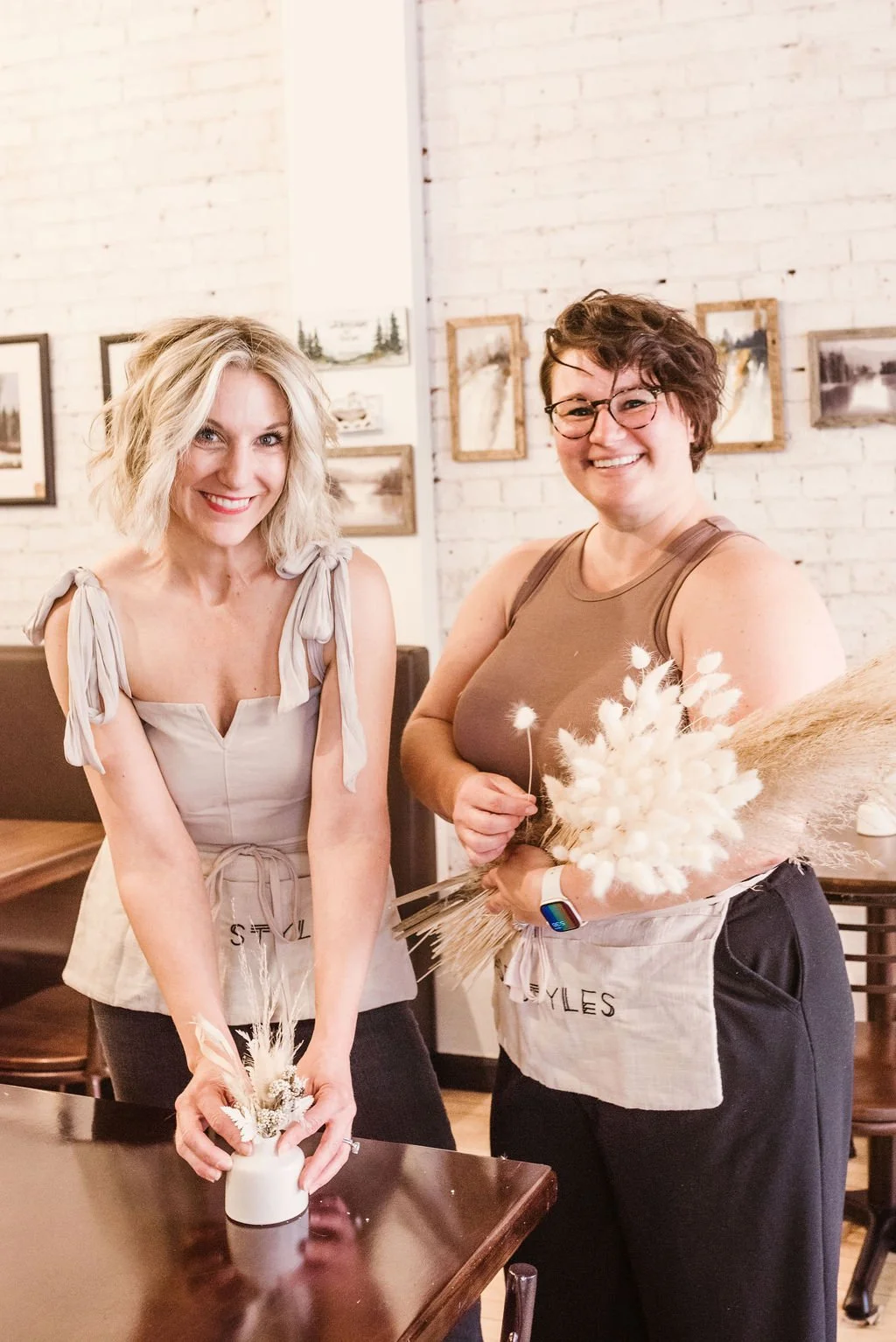 Two smiling women standing indoors, holding bouquets of dried flowers, in a room with white brick walls and framed artwork.