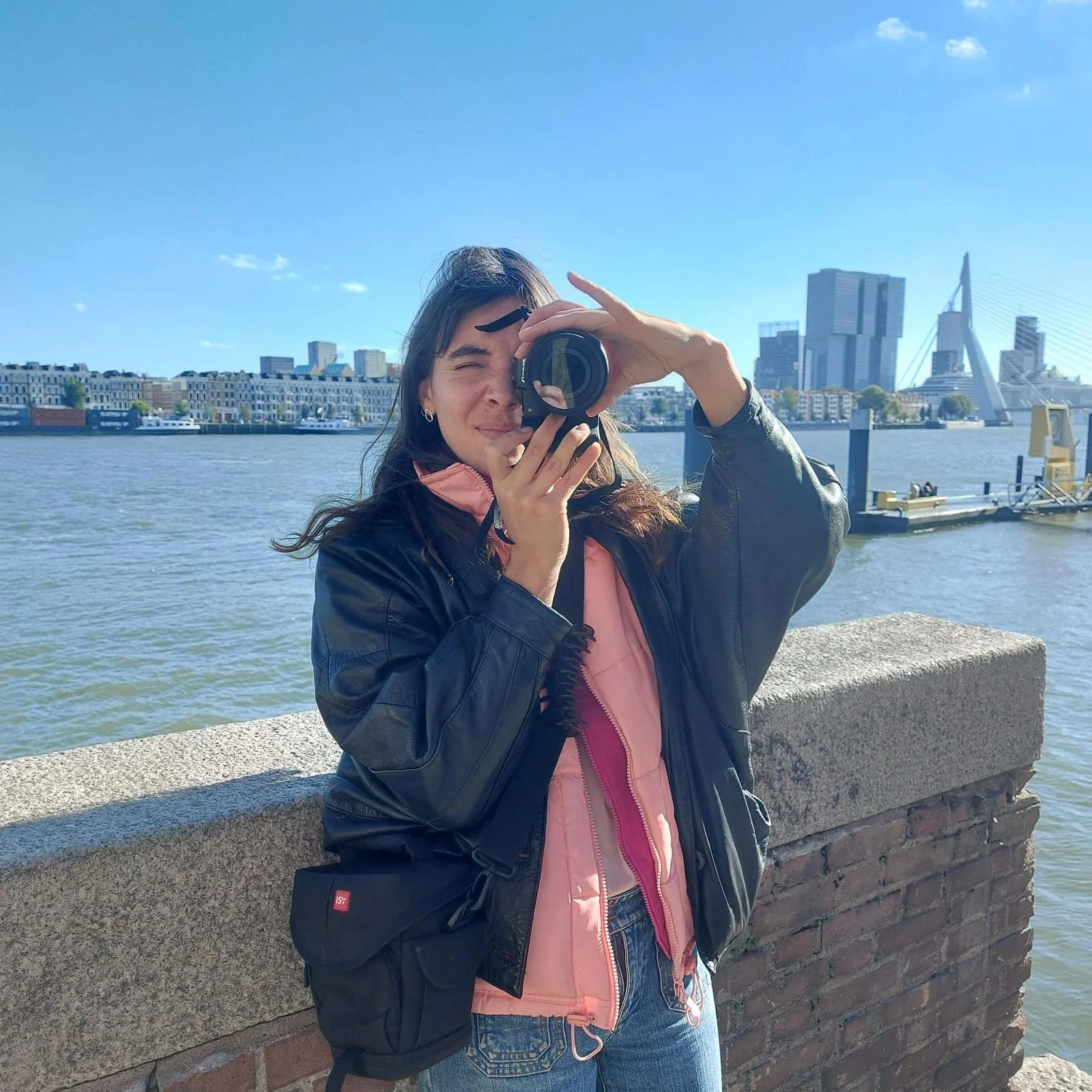 A woman taking a selfie with a river, modern city buildings, and a bridge in the background on a sunny day.