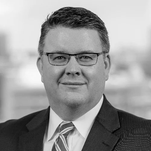 Black and white professional headshot of a middle-aged public affairs consultant with short hair, glasses, wearing a suit and tie, smiling at the camera.