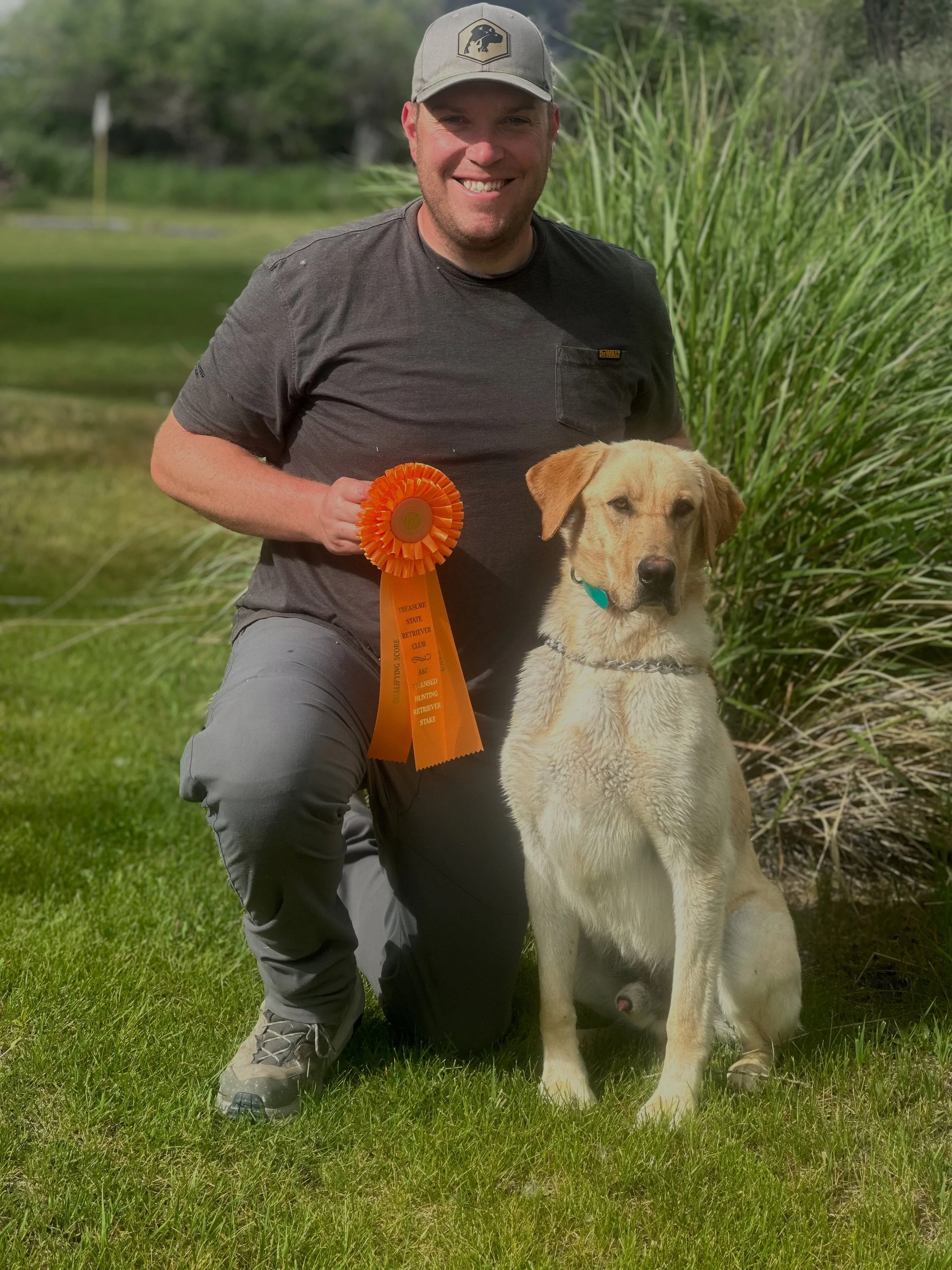 A man kneeling on green grass next to a yellow Labrador Retriever dog, holding an orange award ribbon, outdoors near a pond with plants and trees in the background.