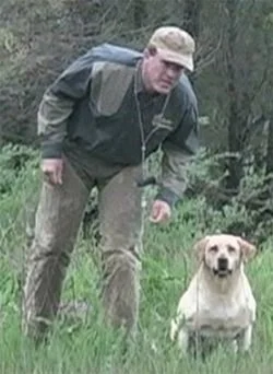 A man in outdoor clothing crouching next to a yellow Labrador retriever in a grassy outdoor area.