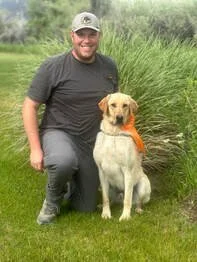 A man with a beard and baseball cap kneels next to a yellow Labrador Retriever wearing an orange bandana in a grassy outdoor area.