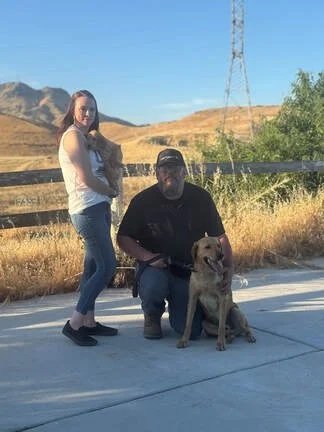 A woman, a man, and a dog outdoors near a fence with hills and a utility pole in the background.