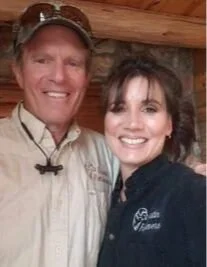 A man and woman smiling, standing close together indoors. The man is wearing a beige shirt and a hat, and the woman has dark hair and is wearing a black jacket.