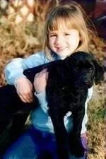 A young girl with blonde hair smiling and holding a black puppy outdoors in a natural setting.