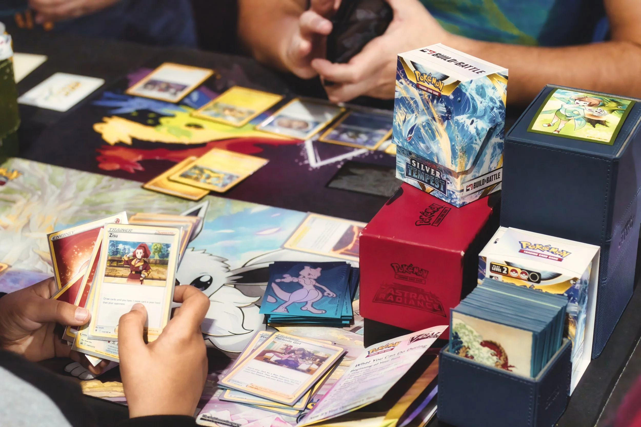 A person is playing a Pokémon trading card game, holding several Pokémon cards in their hands, with various Pokémon cards and booster packs on the table, including a visible booster box labeled 'Silver Tempest' and another box labeled 'Astral Radiance'.