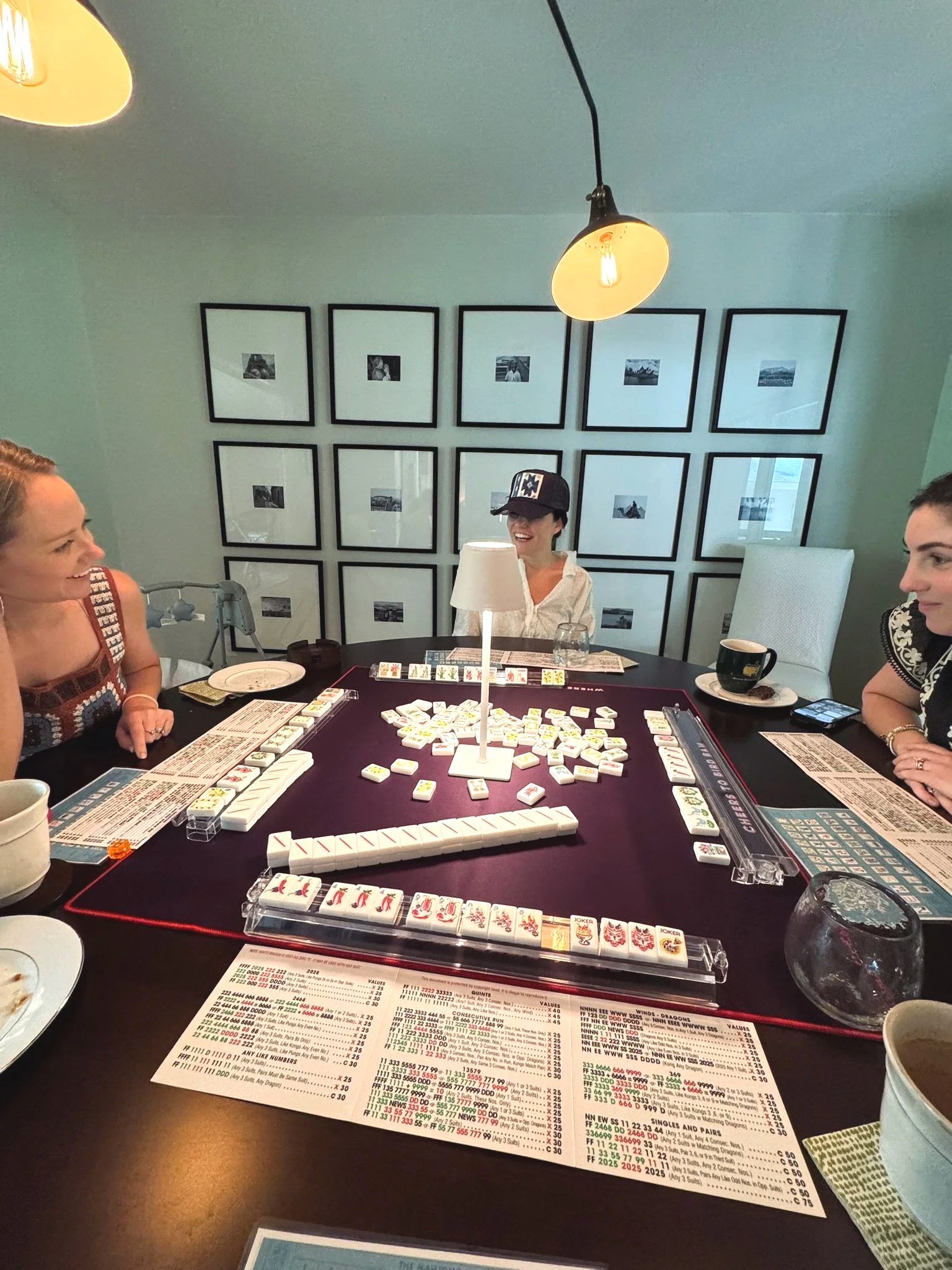 People playing Mahjong around a table with game tiles, score sheets, and drinks in a room with framed photos on the wall and ceiling lights.