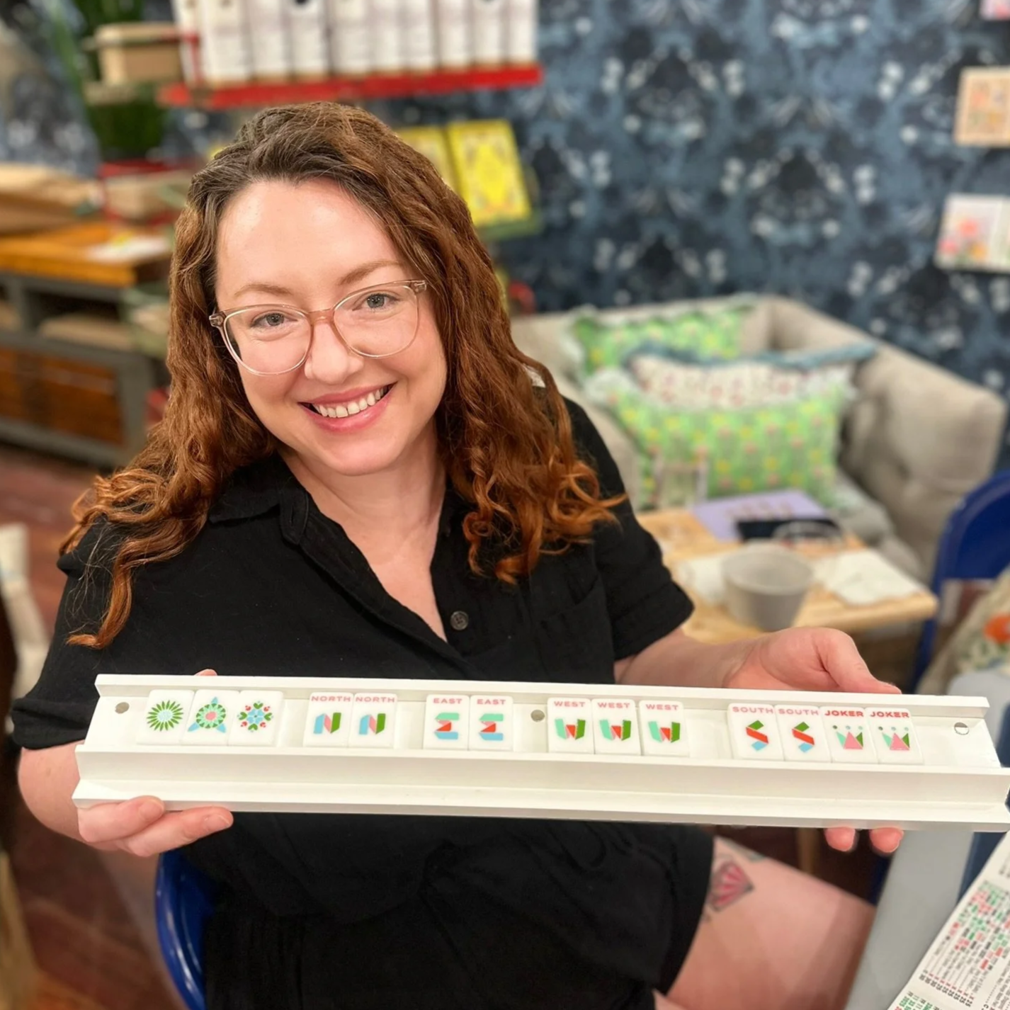 A woman with glasses and curly red hair smiling while holding a tile set of colorful dominoes with directional patterns at a table in a cozy, decorated room.
