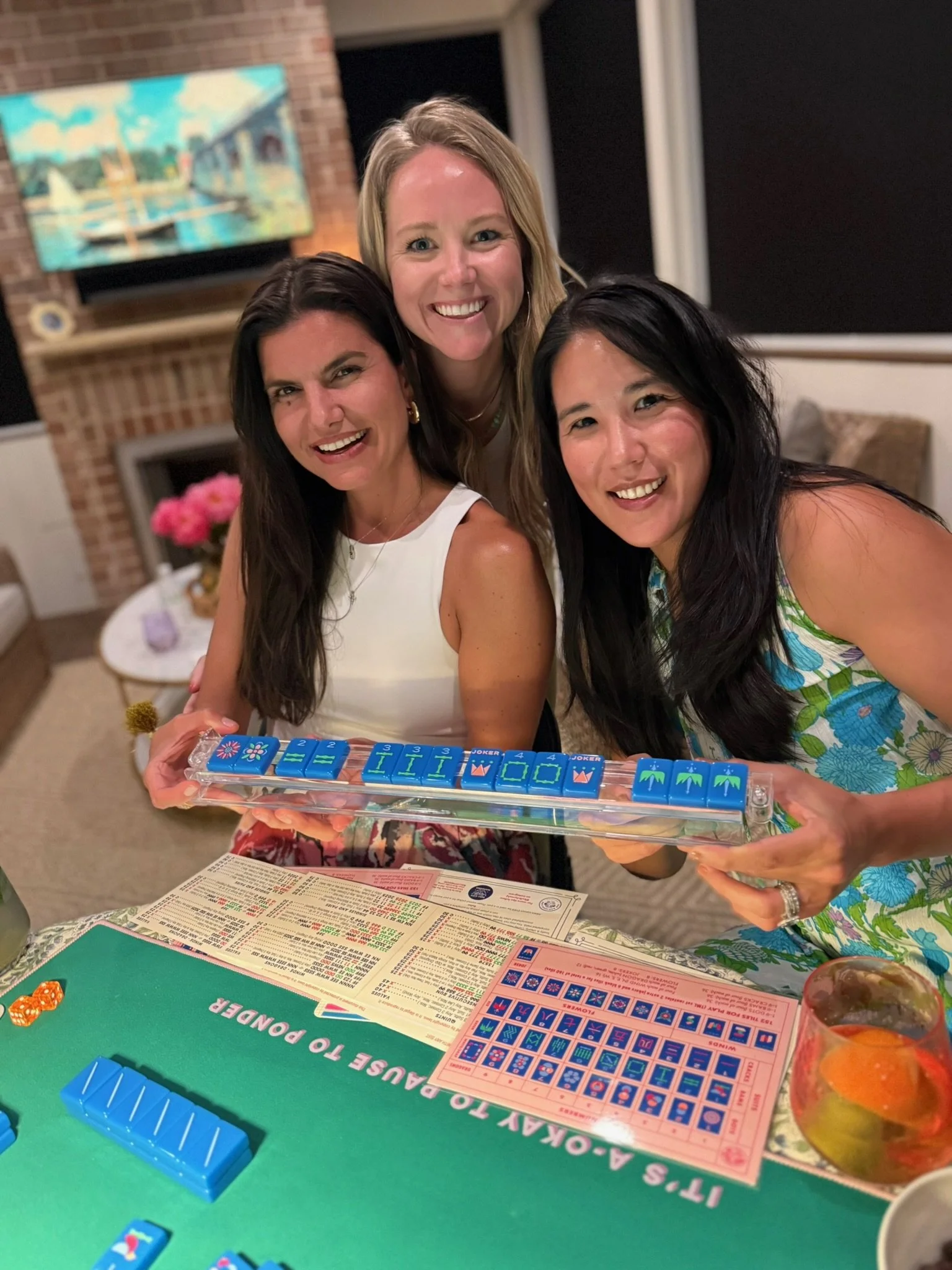 Three women are smiling and posing for a photo around a mahjong game at a table. The table has mahjong tiles, dice, and game cards on it. One woman is holding the mahjong game tray. Behind them is a brick wall with a colorful painting and a window with black blinds. A jar of mixed fruit and a drink are visible on the table.
