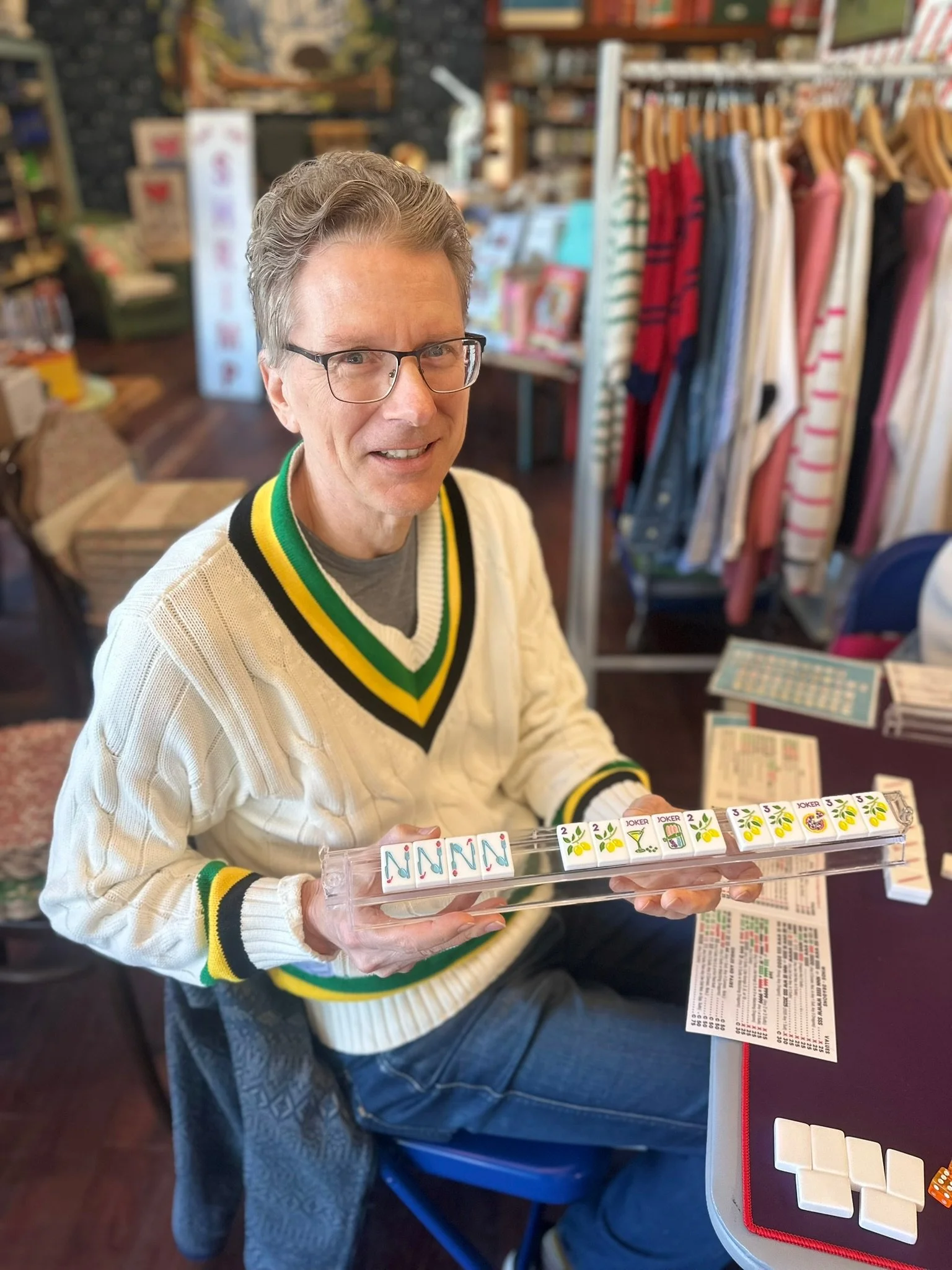 A man with glasses and a beige sweater with green and yellow stripes is holding a transparent rack of domino tiles, smiling at the camera. The domino tiles feature colorful designs, including flowers, cocktails, and the word "JOKER." In the background, there are shelves with books or games, and a table with papers and other game pieces.