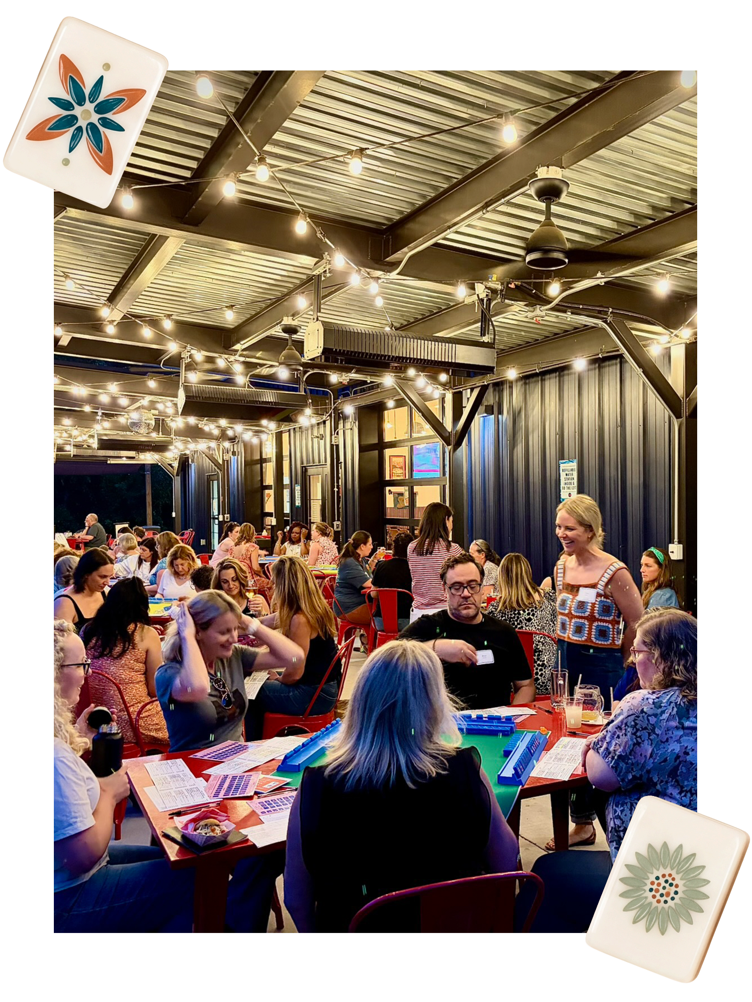 People playing a tabletop mahjong board game at a large indoor gathering with string lights and metallic ceiling, some are sitting and others are standing, with colorful game pieces and papers on the tables.