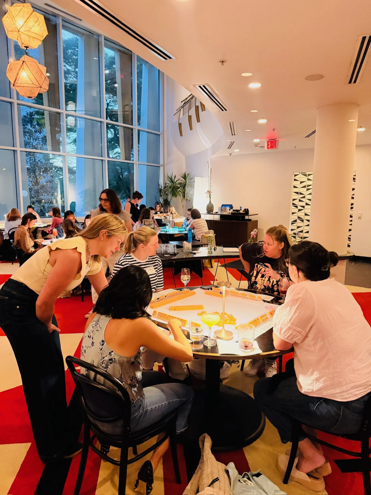 People playing a mahjong game at a round table in a brightly lit, modern restaurant with large windows and hanging lights.