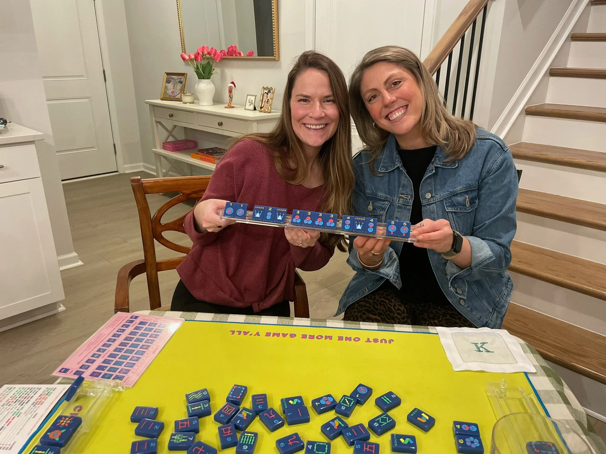 Two women smiling and holding a domino tile with colorful symbols at a table with a yellow game board, in a cozy indoor setting with decorated shelves and staircase in the background.
