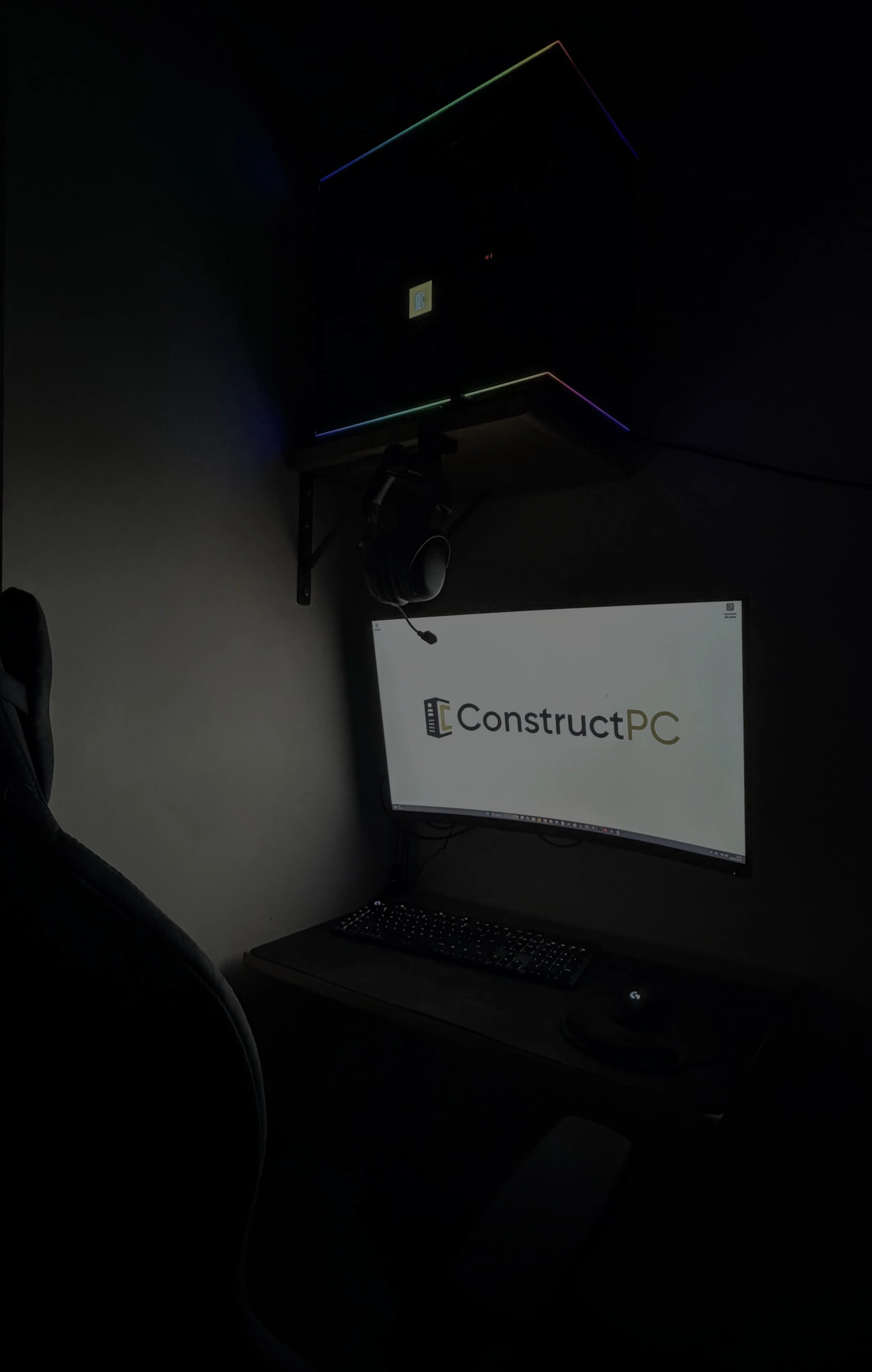 Computer setup in a dark room with a monitor displaying the ConstructPC logo, a keyboard, and a headset hanging on a wall-mounted shelf.
