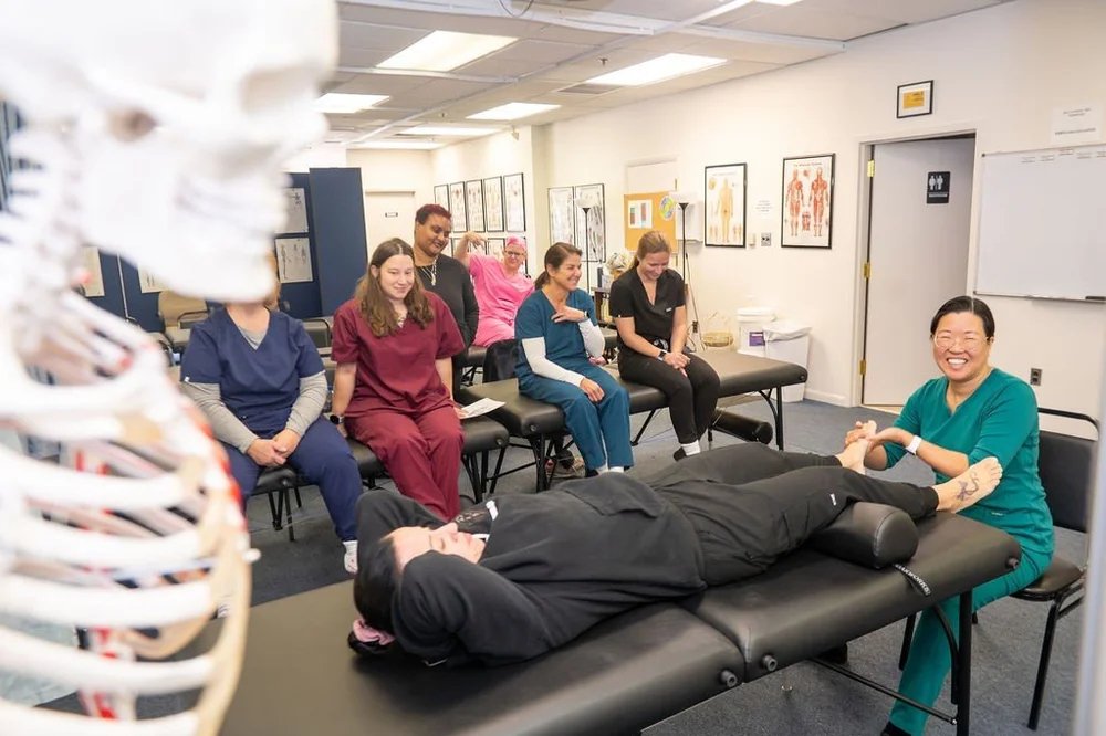 A group of healthcare professionals in scrubs and uniforms watching a demonstration at a medical training session, with a healthcare worker practicing a procedure on a patient lying on a treatment table.