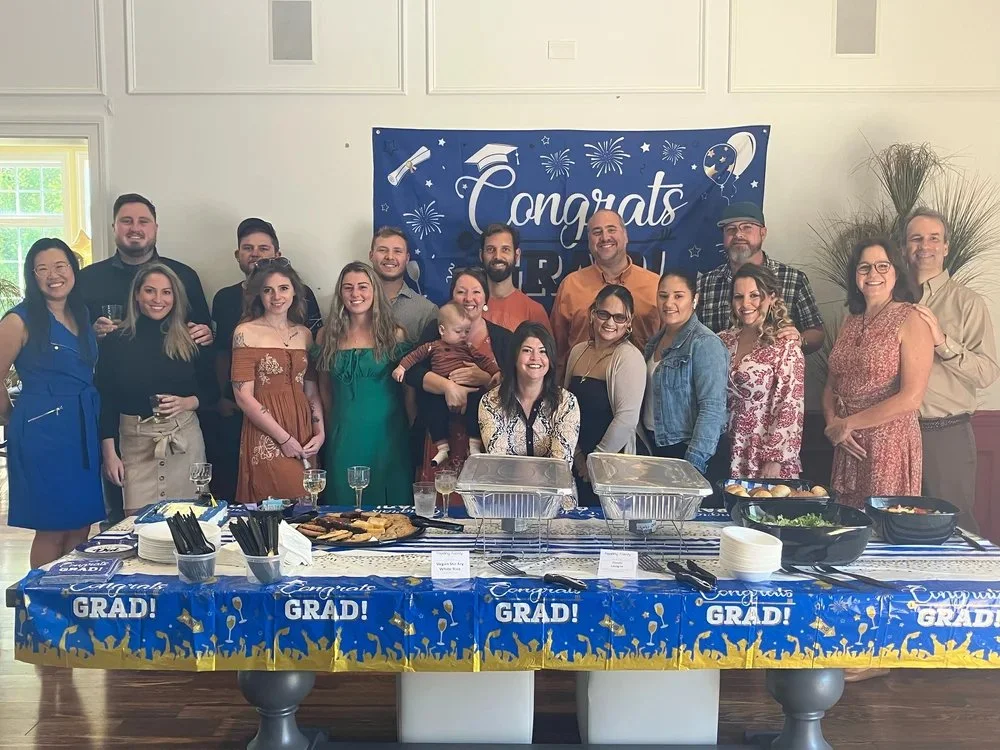 Group of people celebrating graduation at a party with a decorated table and a blue banner that says 'Congrats Grad' in a hall with white walls and windows.