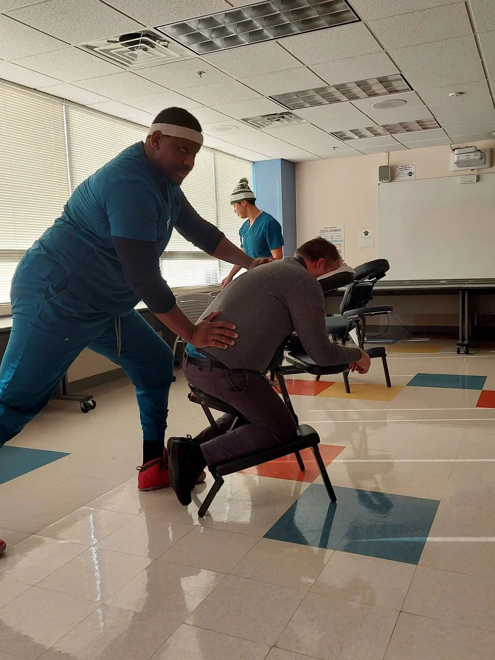 A man receiving back massage from a healthcare professional while seated and leaning forward in a chair in a room with bright windows and colorful floor tiles.