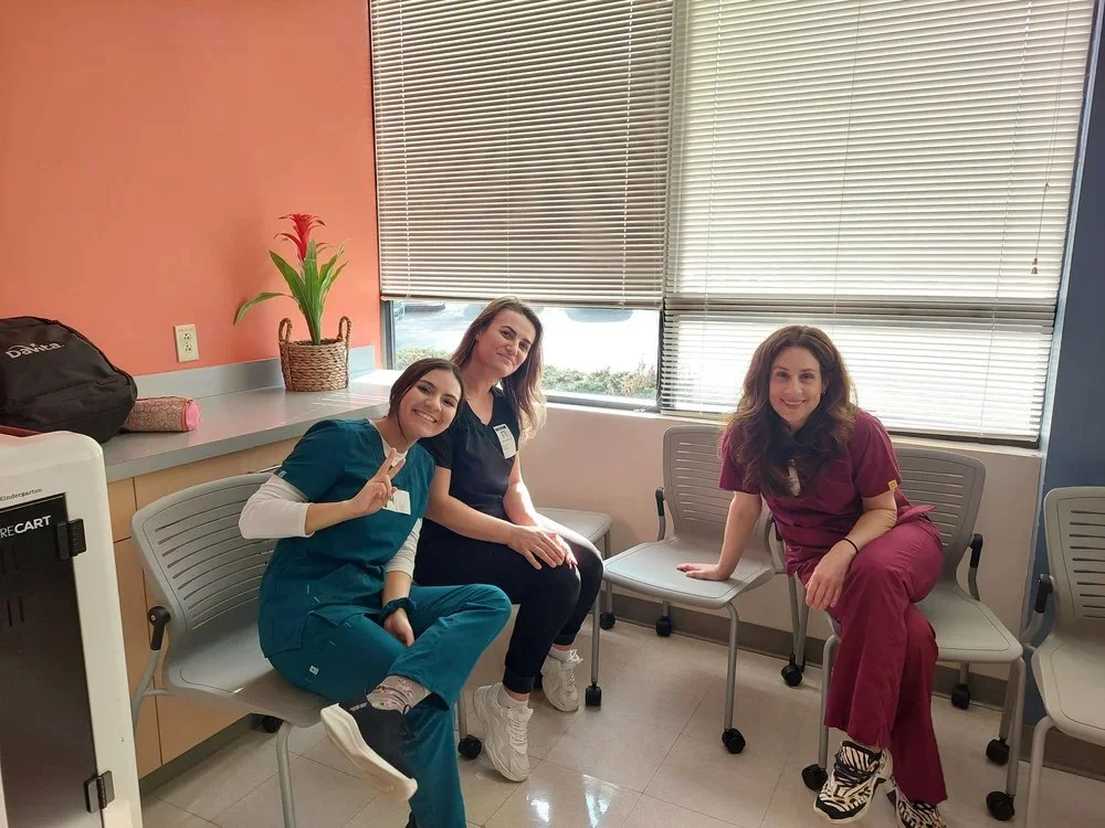 Three women sitting on chairs in a room, two in medical scrubs and one in casual clothes, smiling at the camera.