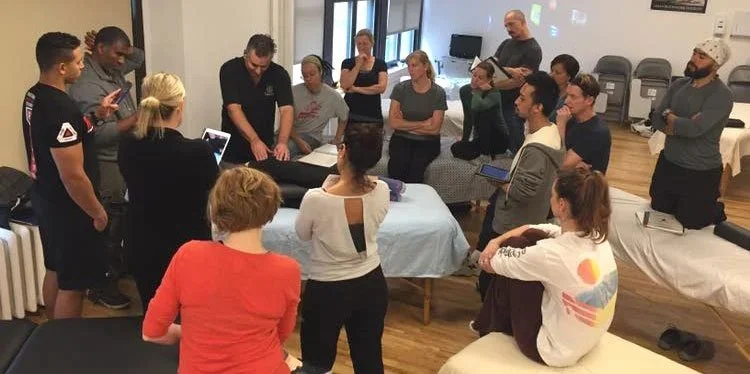 A group of people gathered in a room, standing and sitting around a table with a person demonstrating or explaining something, while others listen and observe. Some are using tablets or phones.