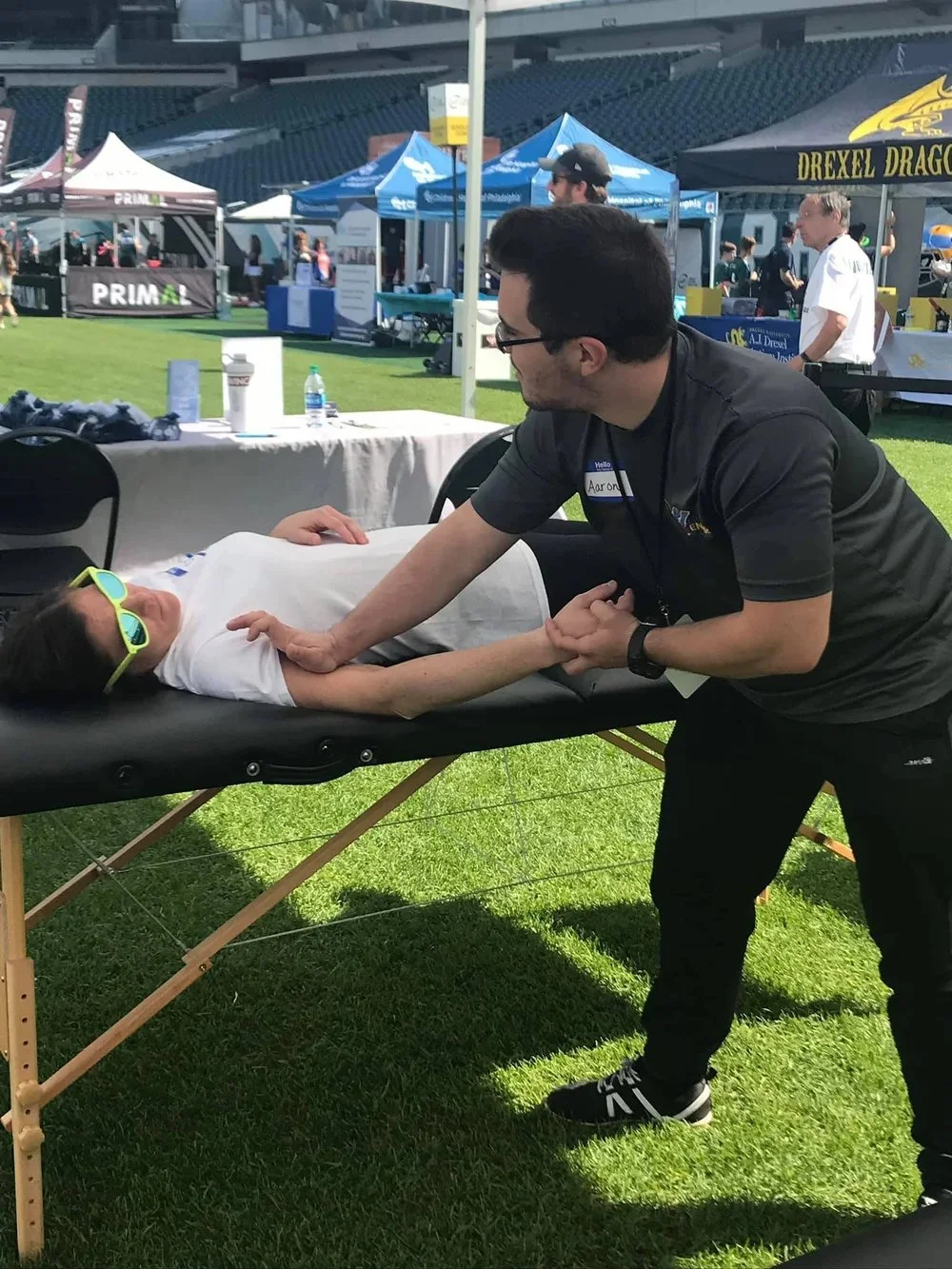 A young girl lying on a massage table outdoors, wearing sunglasses, with a man leaning over her holding her hand. The scene is at an outdoor event with tents and people in the background.