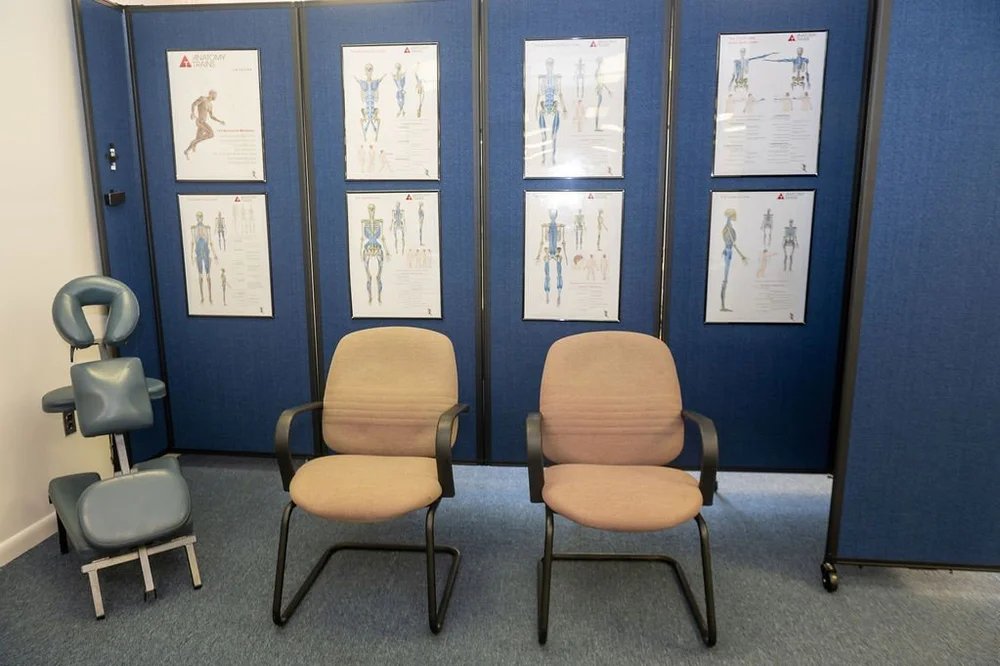 Office or clinical waiting room with two beige chairs, a backrest and armrest, and a chiropractic or medical post in the background with posters of human anatomy and skeletal diagrams, and a gray back/neck support cushion.