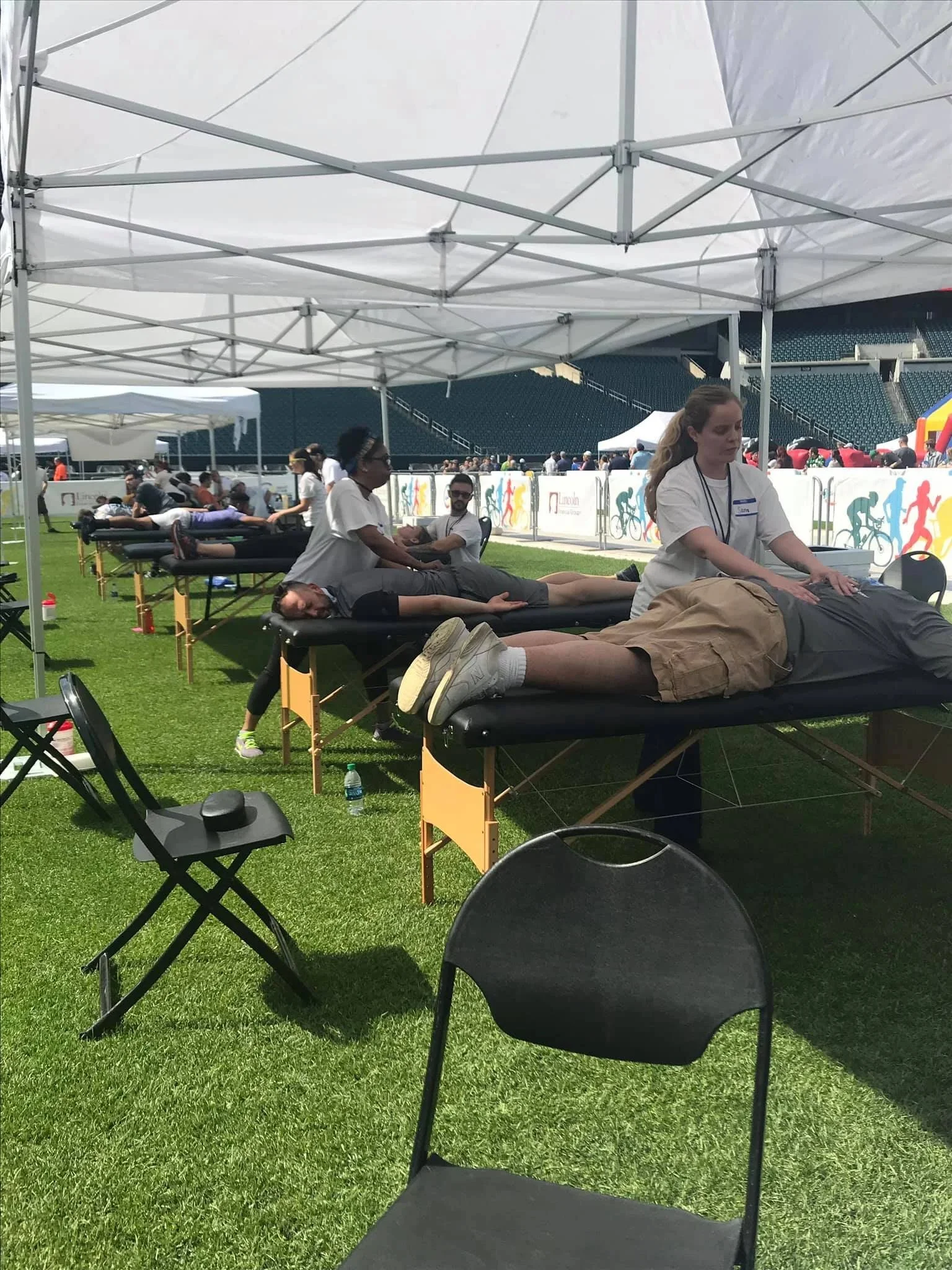 People receiving massages under white canopy tents on a grassy field at an outdoor event.