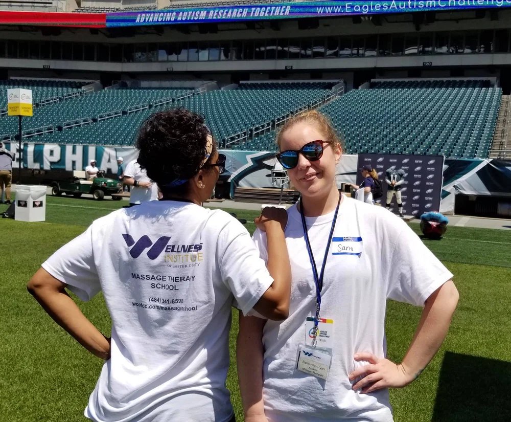 Two women stand on a football field during an autism awareness event, with empty stadium seats in the background. One woman faces away from the camera, wearing a white t-shirt with the word "Wellness" and contact information on the back. The other woman faces the camera, wearing sunglasses, a white t-shirt with a name tag that says "Sam," and a lanyard. They are smiling and posing together.