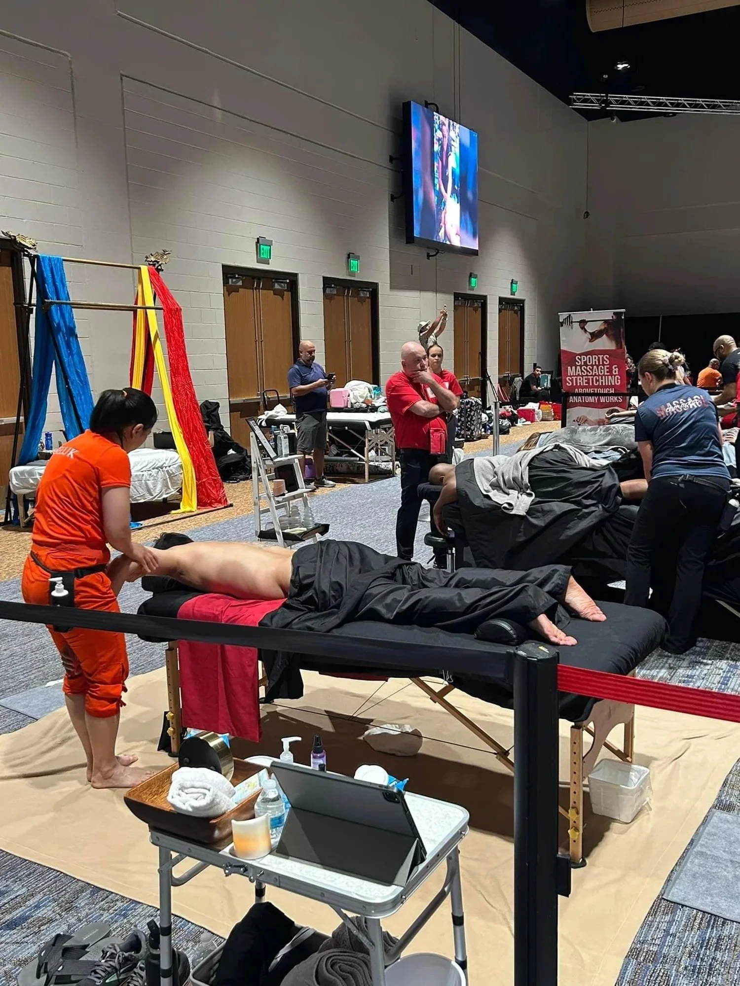 A massage therapist is giving a massage on a massage table at an indoor event with several people nearby. There are medical supplies, water bottles, and towels around the massage area. Multiple people are observing or involved, including a woman in a