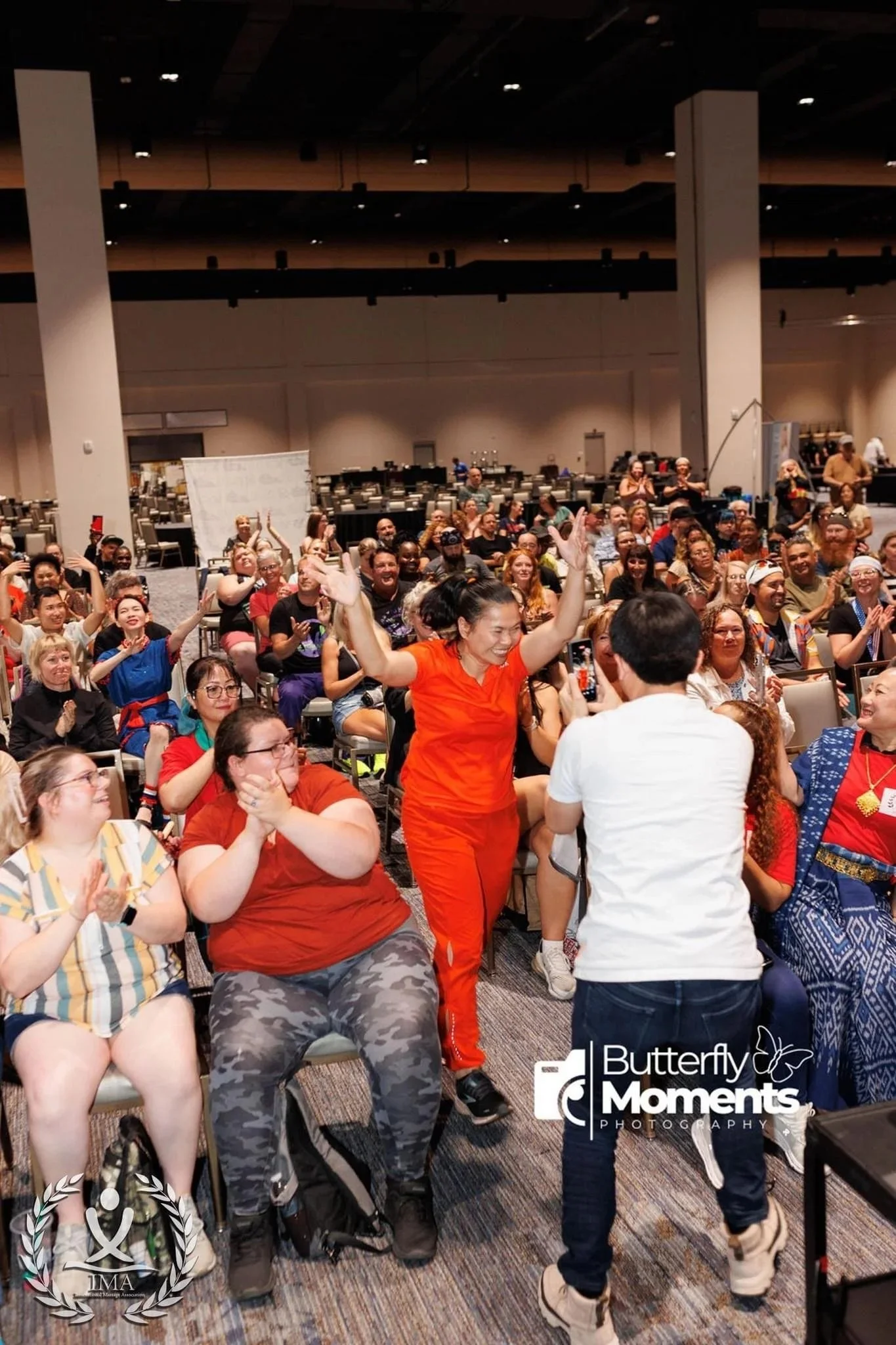 A group of diverse people sitting and standing in a conference room, smiling and enjoying a lively event with someone taking a photo in the foreground.
