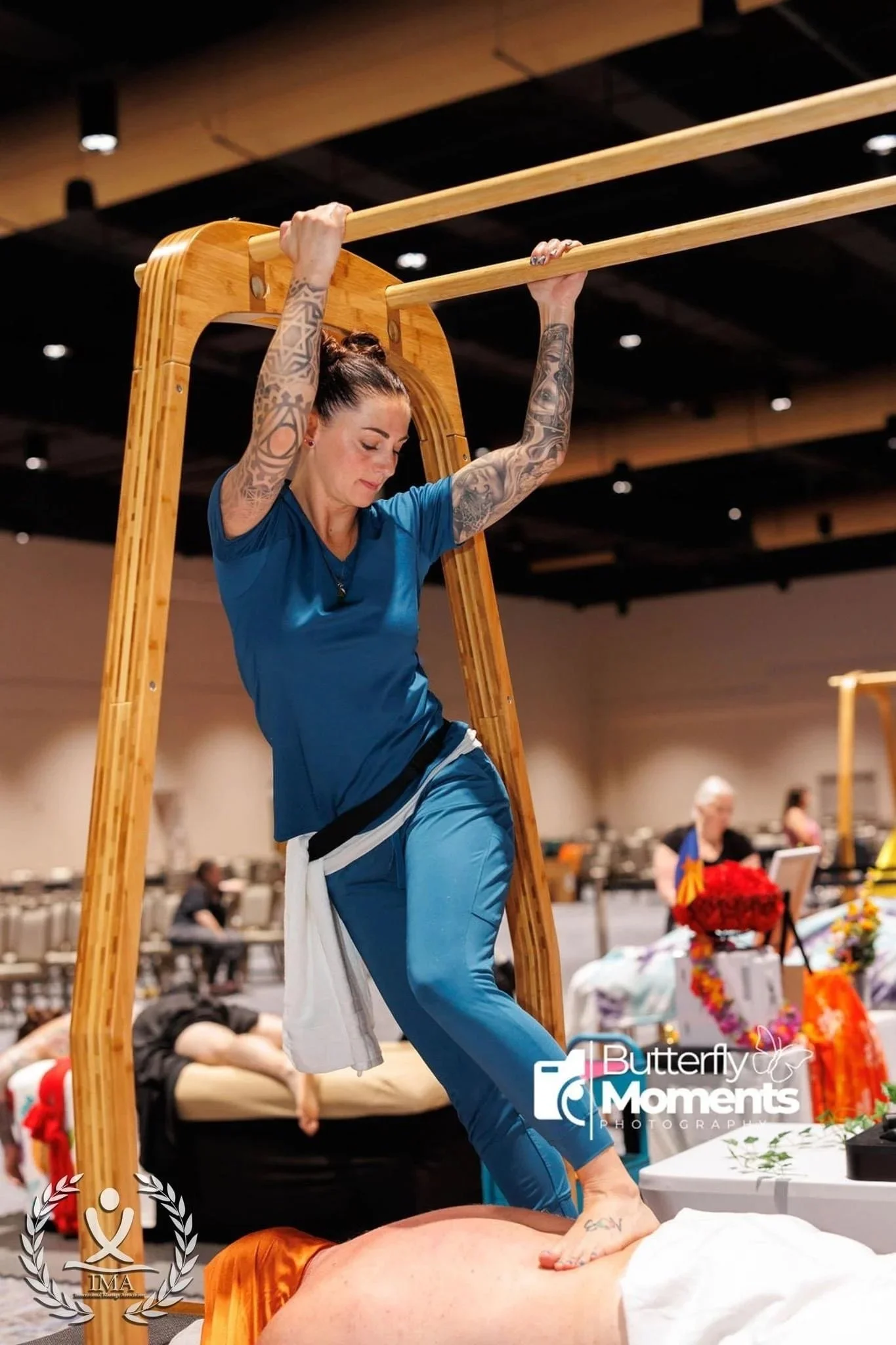 A woman with tattoos on her arms performing massage or healing on a man lying face down on a table at an indoor event with tables and people in the background.