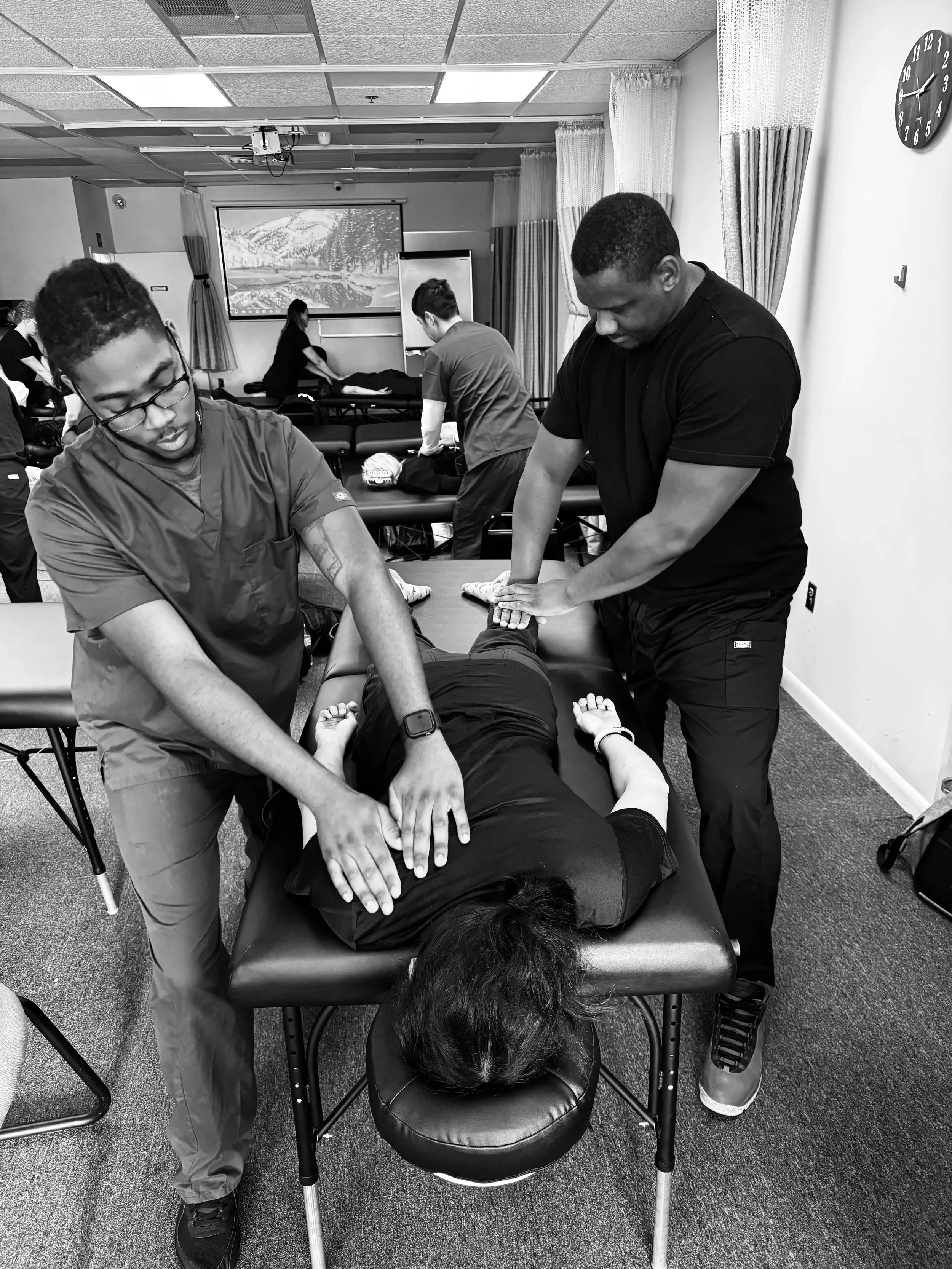 A group of healthcare professionals performing CPR on a person lying face down on a massage table in a training room. Others are preparing or practicing in the background.