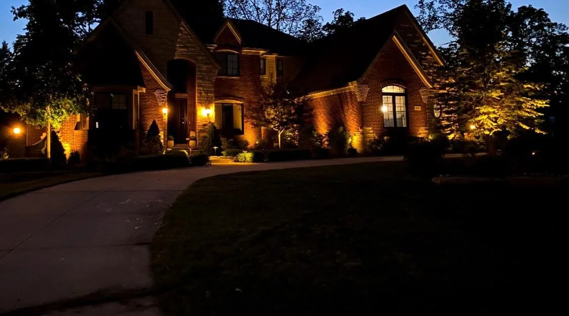 A large brick house at night with exterior lights illuminating the front entrance and surrounding trees, a curved driveway leading up to it, and a dark sky overhead.